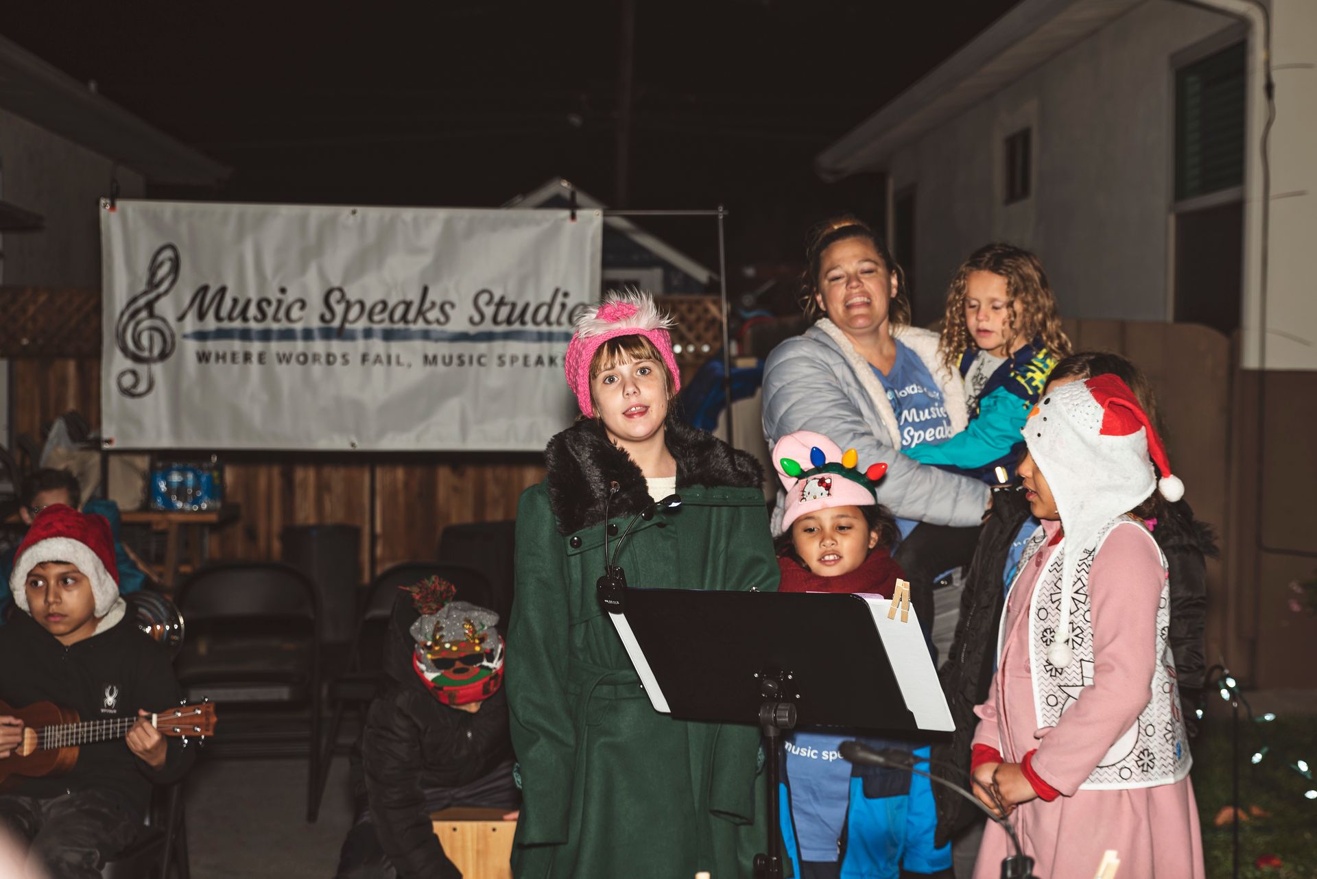 A group of children are singing christmas carols in front of a sign that says music speaks studio.