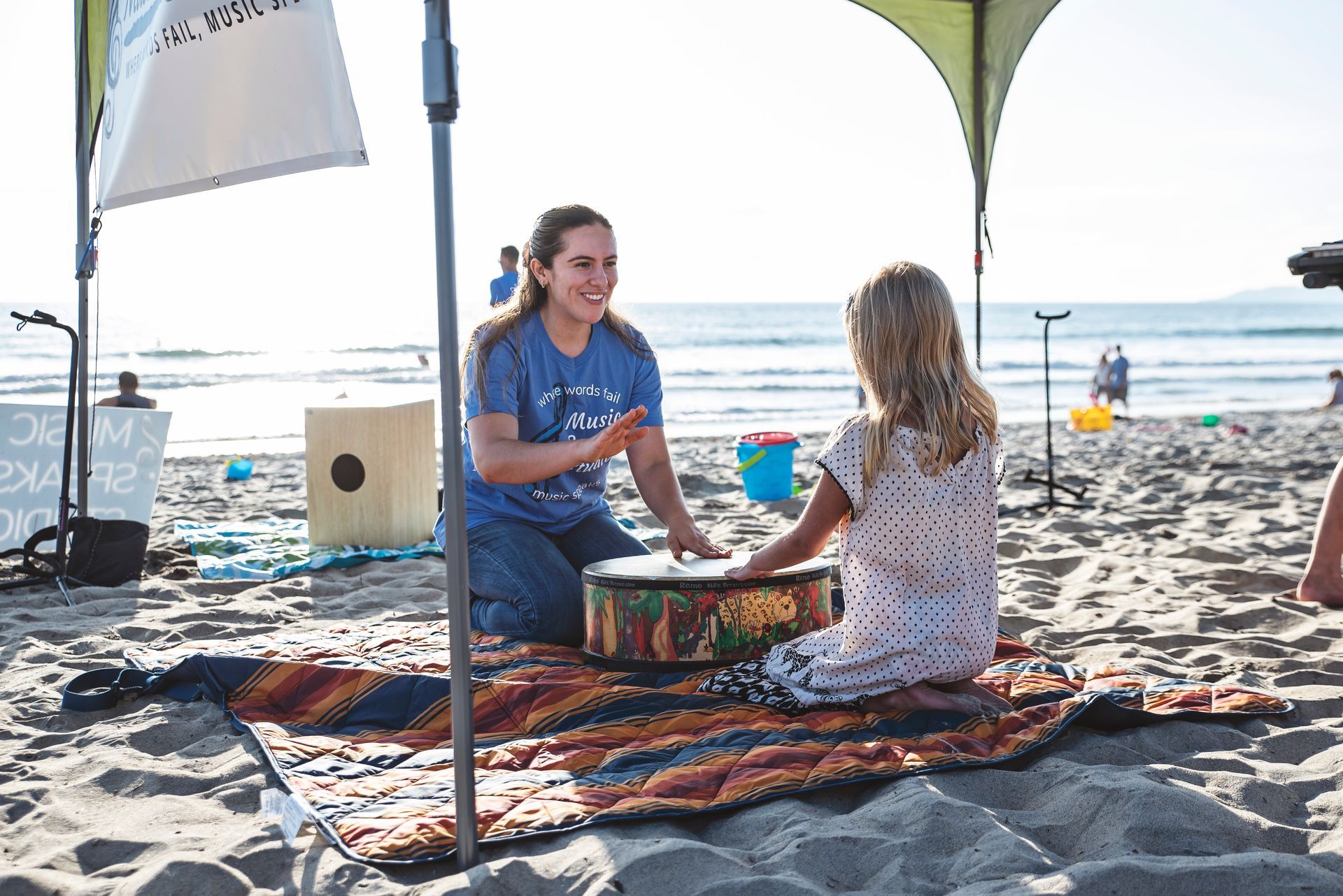 A woman is teaching a little girl how to play a drum on the beach.