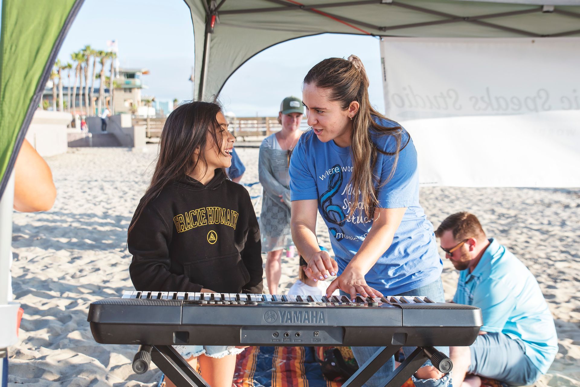 A woman is teaching another woman how to play a keyboard on the beach.