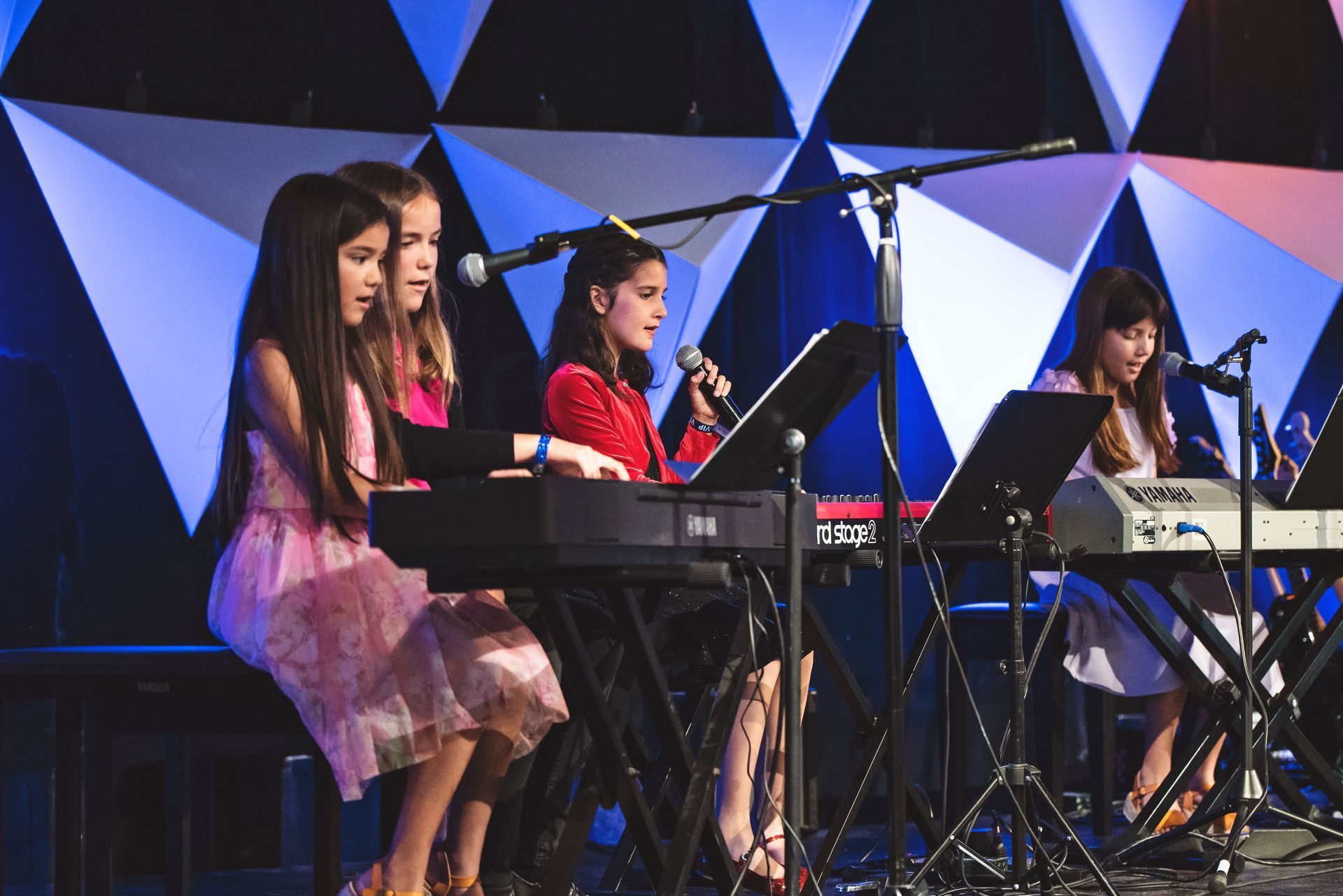 A group of young girls are playing keyboards and singing into microphones on a stage.