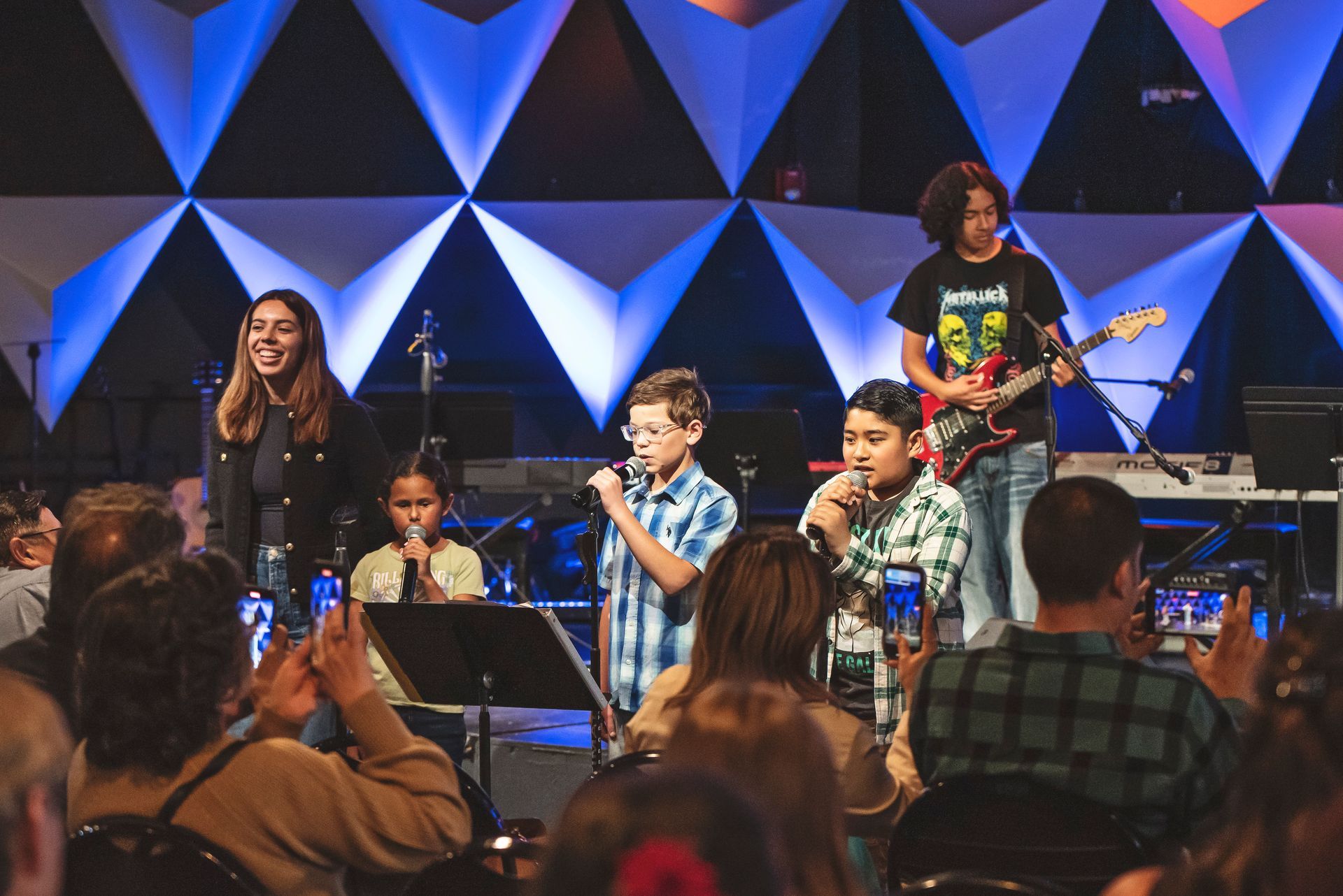 A group of children are singing and playing instruments on a stage in front of a crowd.