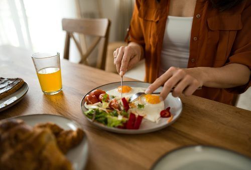 A person in a rust-colored shirt cuts fried eggs and a salad on a wooden table with a glass of orange juice nearby.