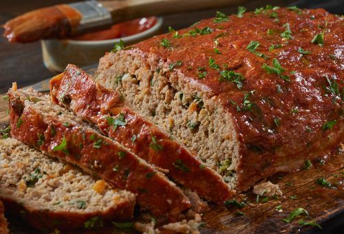 A sliced meatloaf with a glossy red glaze and herbs on top, displayed on a wooden board next to a small dish of sauce.