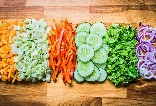Chopped carrots, celery, bell peppers, cucumber slices, lettuce, and red onion rings arranged in rows on a wooden board.