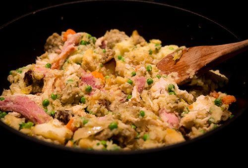 A wooden spoon stirs a savory mixture of cooked chicken, peas, diced carrots, and stuffing in a large pan.