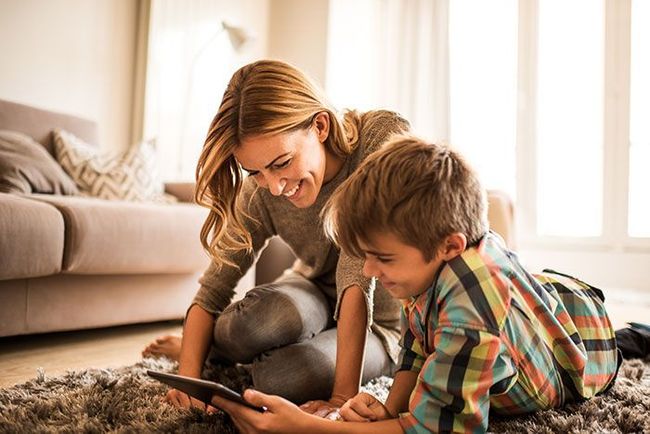 Adult and child kneeling on a rug, smiling while using a tablet in a bright living room.