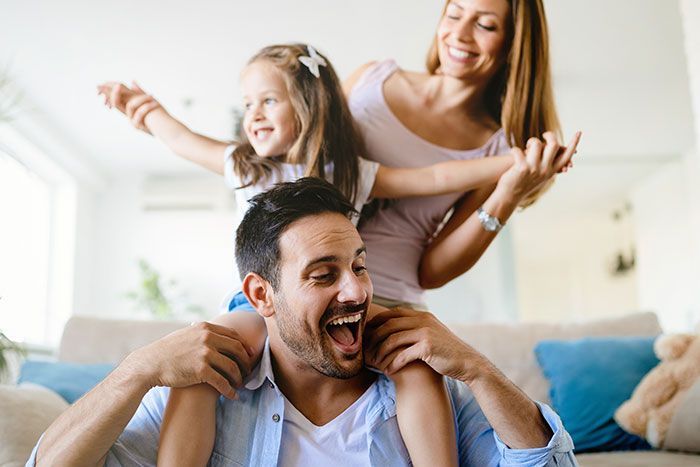 Smiling family playing on a couch in a bright living room, with a child on the father’s shoulders.