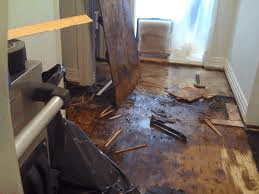 Flood-damaged room with warped hardwood floor, debris, and a chair near a bright window.