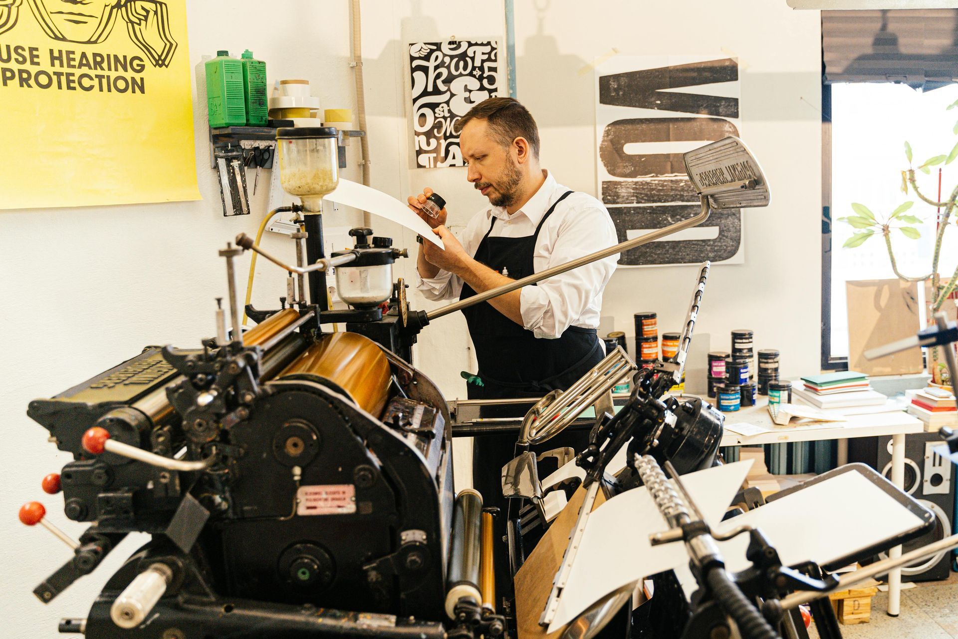 Man in apron examining paper at a printing press in a workshop. Yellow sign and tools are visible.