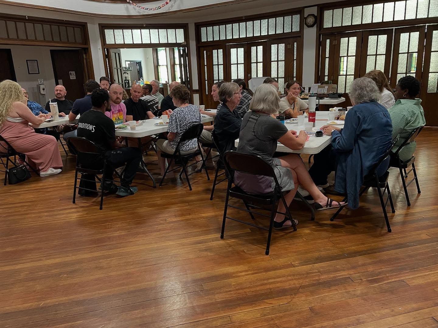 Short North Church. People seated around tables in a bright community hall, eating and talking on a wooden floor.