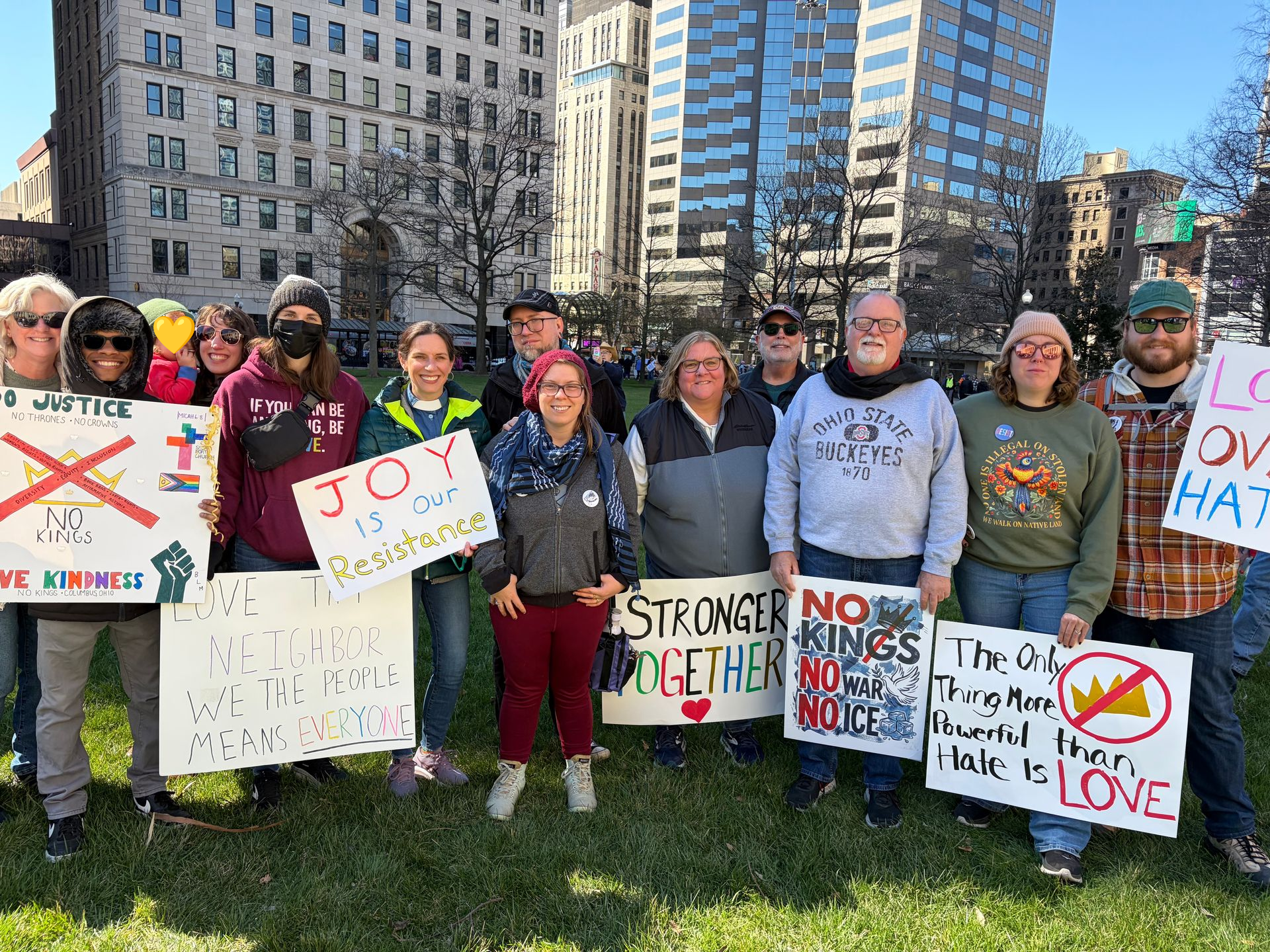 Short North Church. People holding protest signs on a city sidewalk in front of tall buildings, with messages about love and unity.