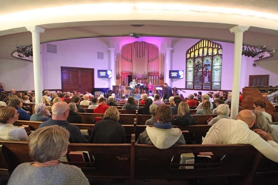 Short North Church. Congregation seated in a church during a service, facing a lit altar with stained-glass windows.