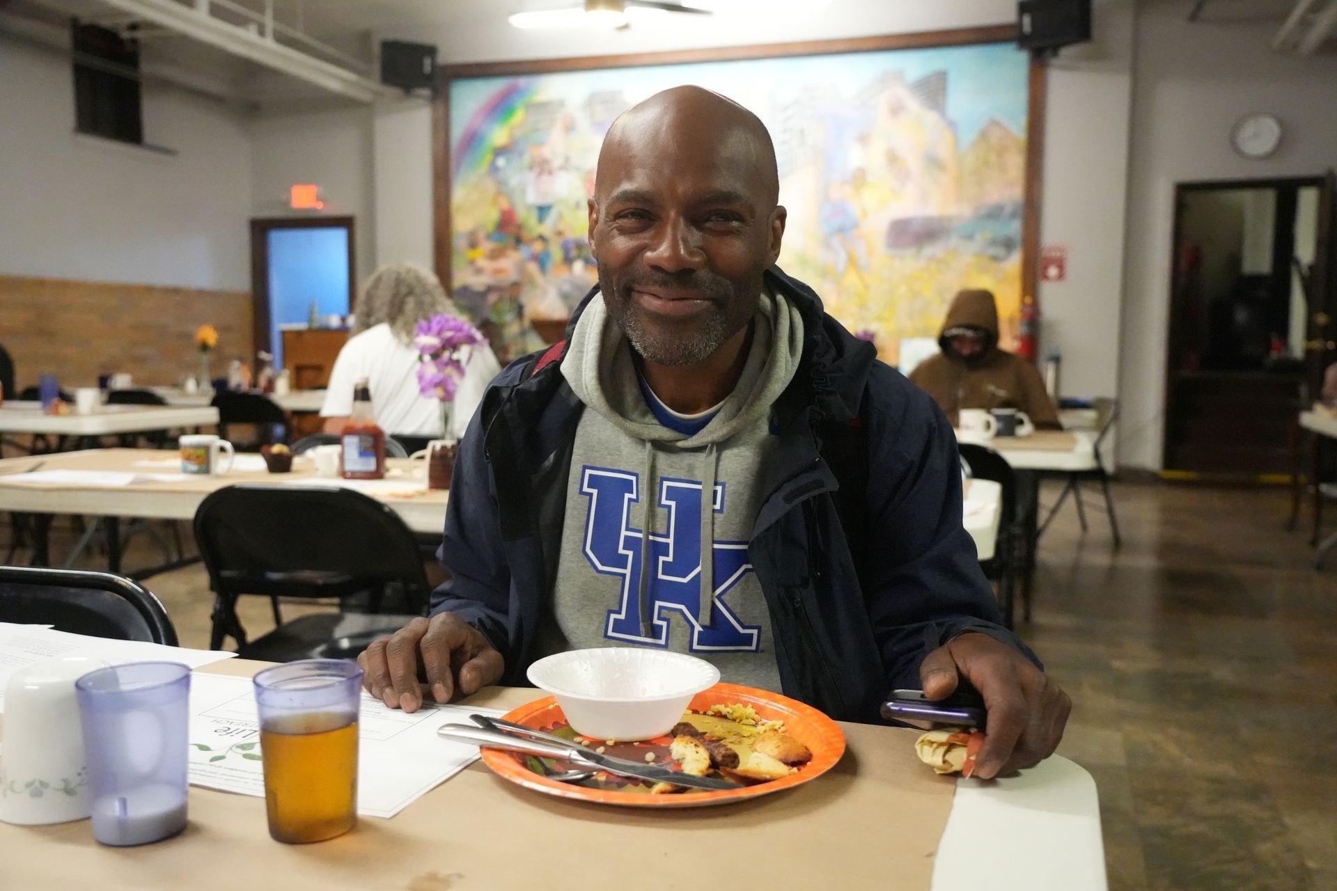 Short North Church. Man smiling at a cafeteria table with a meal, drink, and orange juice in front of a colorful mural.