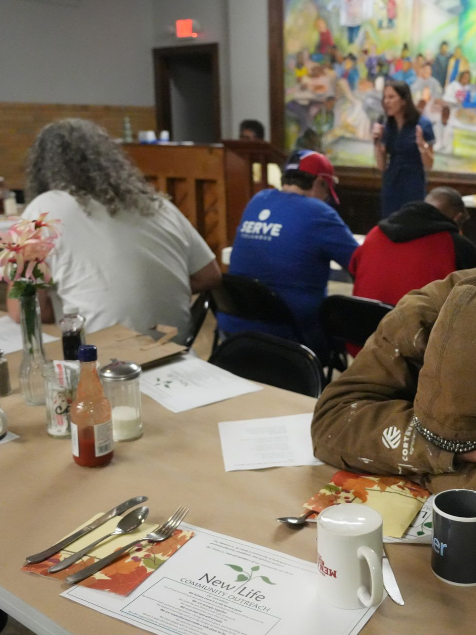 Short North Church. People seated at a banquet table in a hall, with a speaker at the front near a colorful mural.