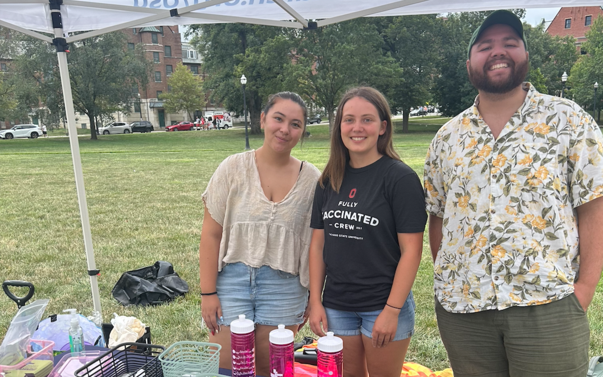 Short North Church at Ohio State. Three people smiling behind a table at an outdoor booth under a white canopy on the Ohio State University oval.