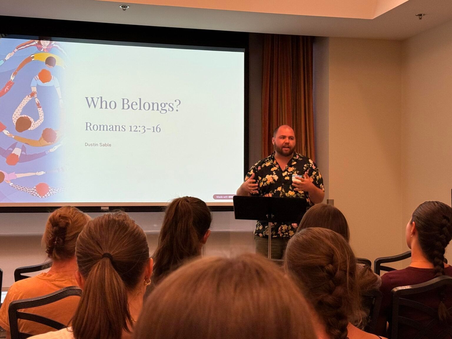 Dustin Sable at podium giving presentation in a conference room, audience seated, slide reads “Who Belongs?”