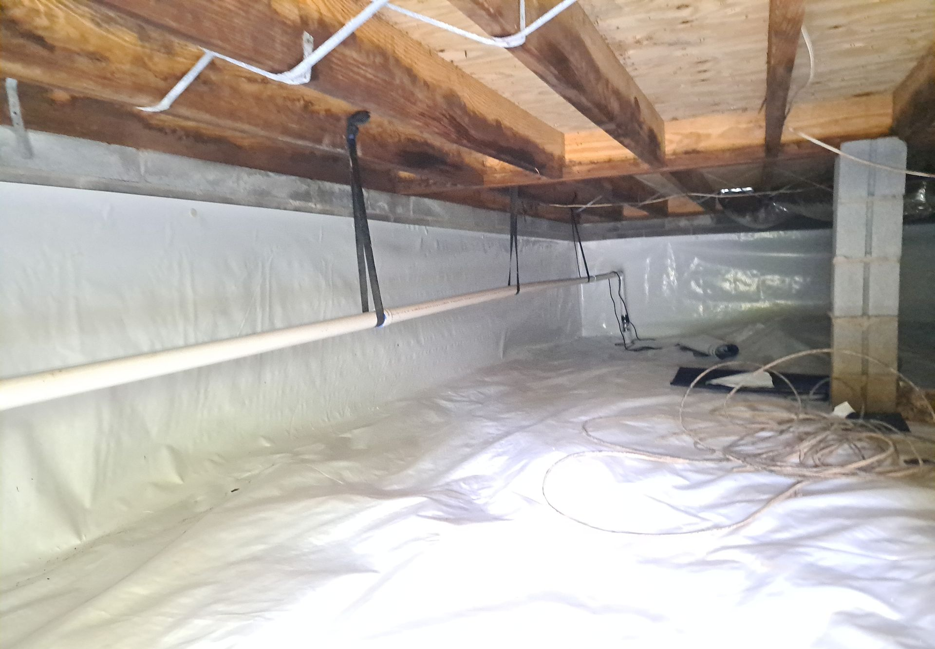 Crawl space with white vapor barrier, wooden beams, support posts, and suspended pipe.
