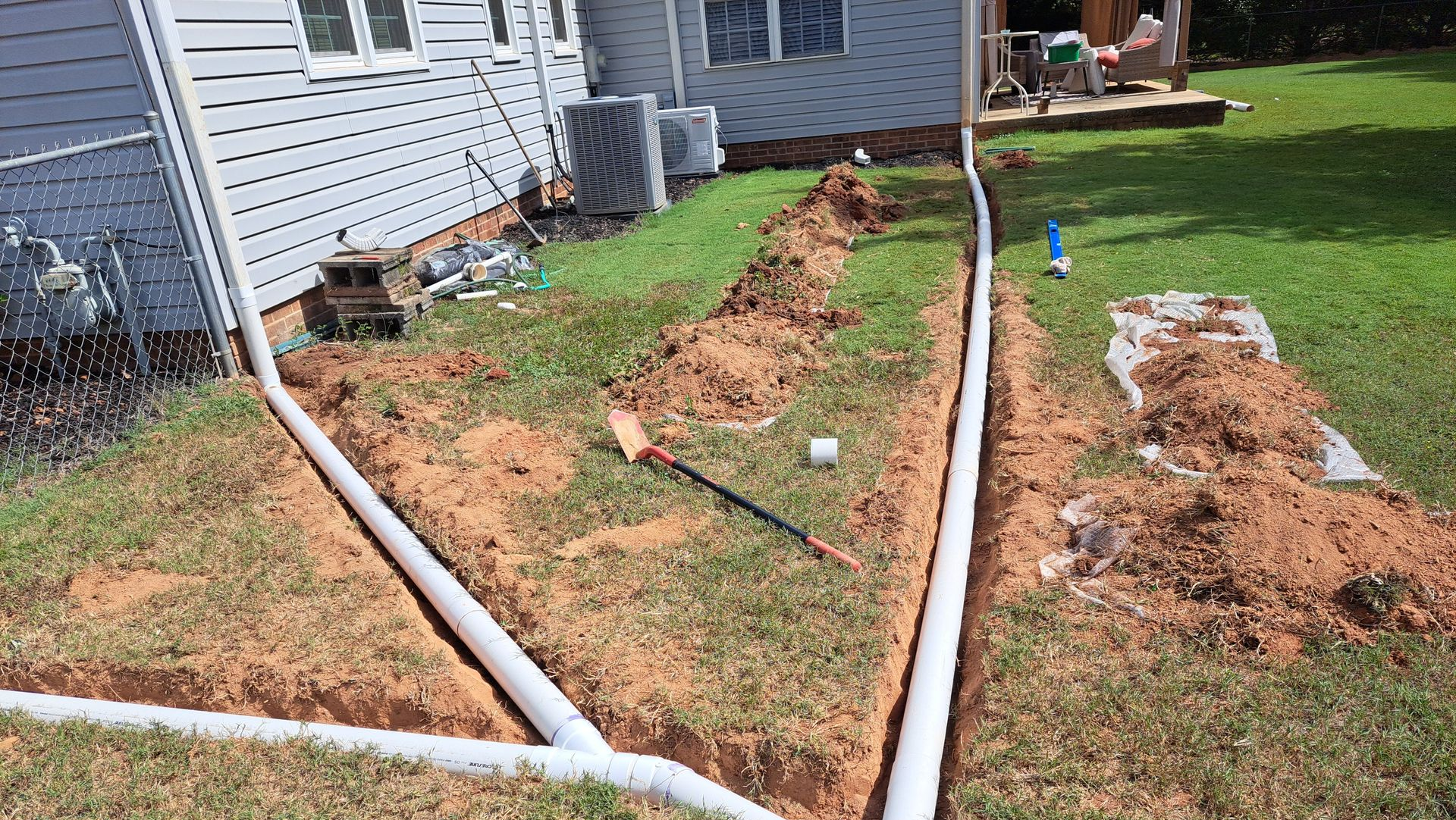 Trenches dug in yard with white pipes installed, likely for drainage near a house.