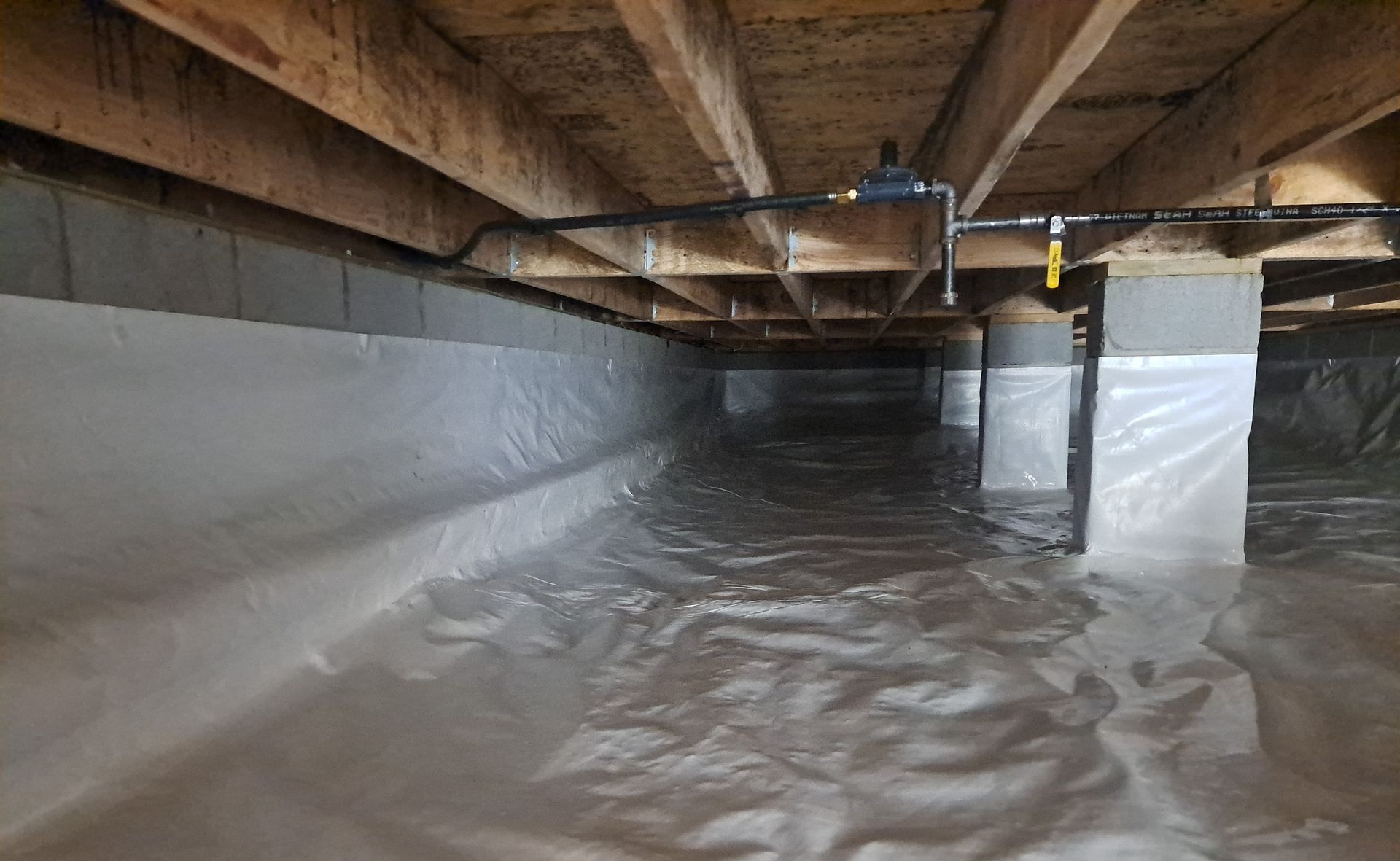 Crawl space with white vapor barrier covering the ground and walls, with wooden beams above.