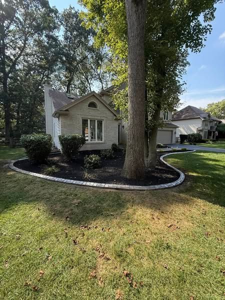 A light brick house with a curved landscaped garden bed featuring dark mulch, shrubs, and a large tree in the front yard.