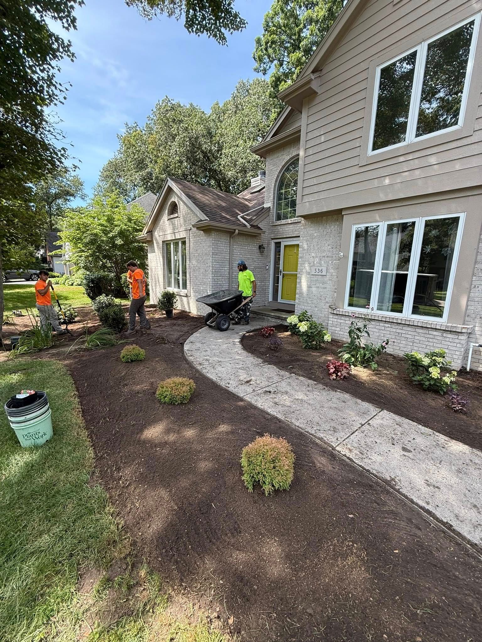 Three workers in neon shirts landscape the front yard of a two-story stone and siding home with a curved stone path.