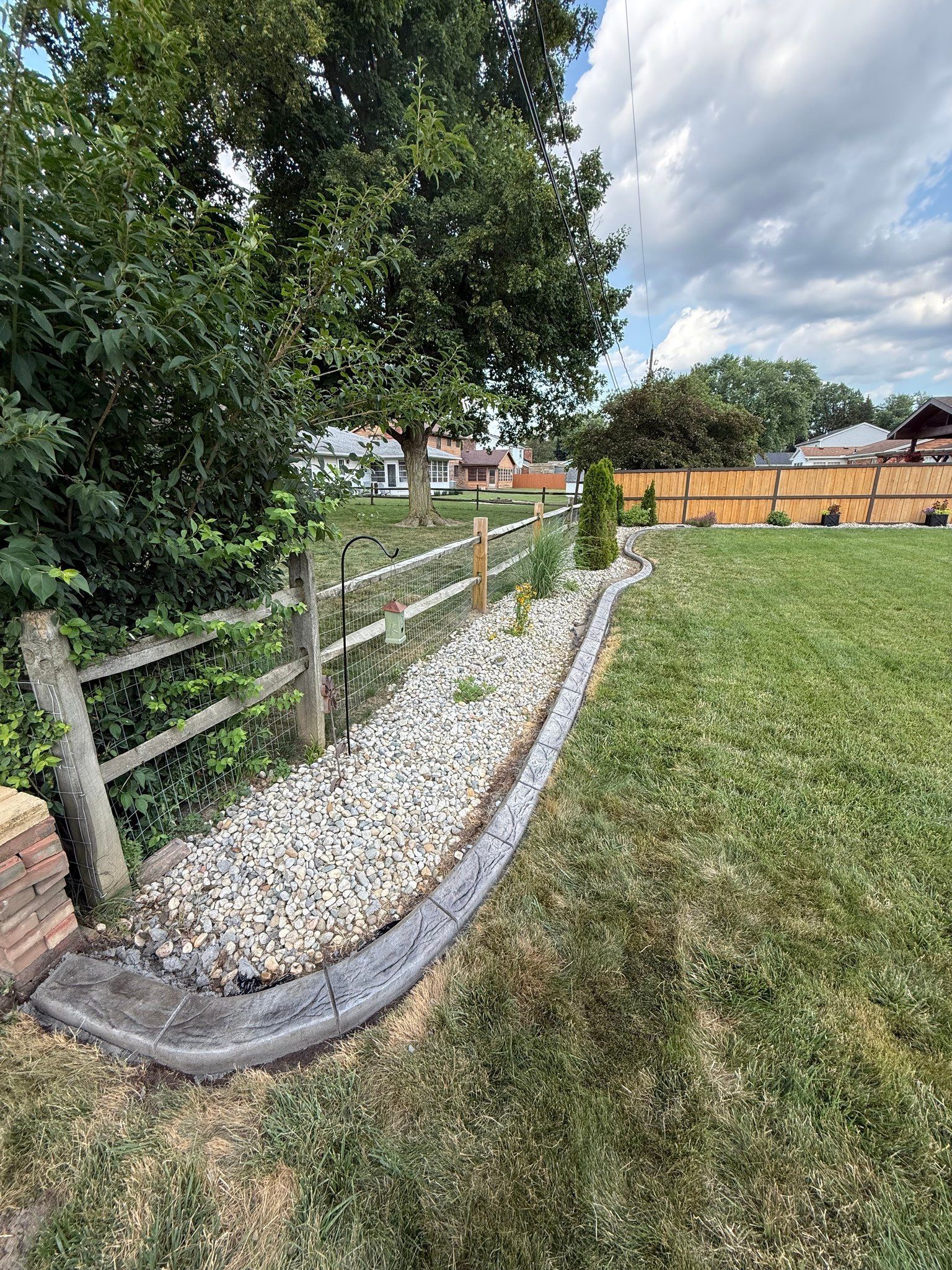A long, curving garden bed filled with light gravel, bordered by decorative concrete edging next to a wooden fence.