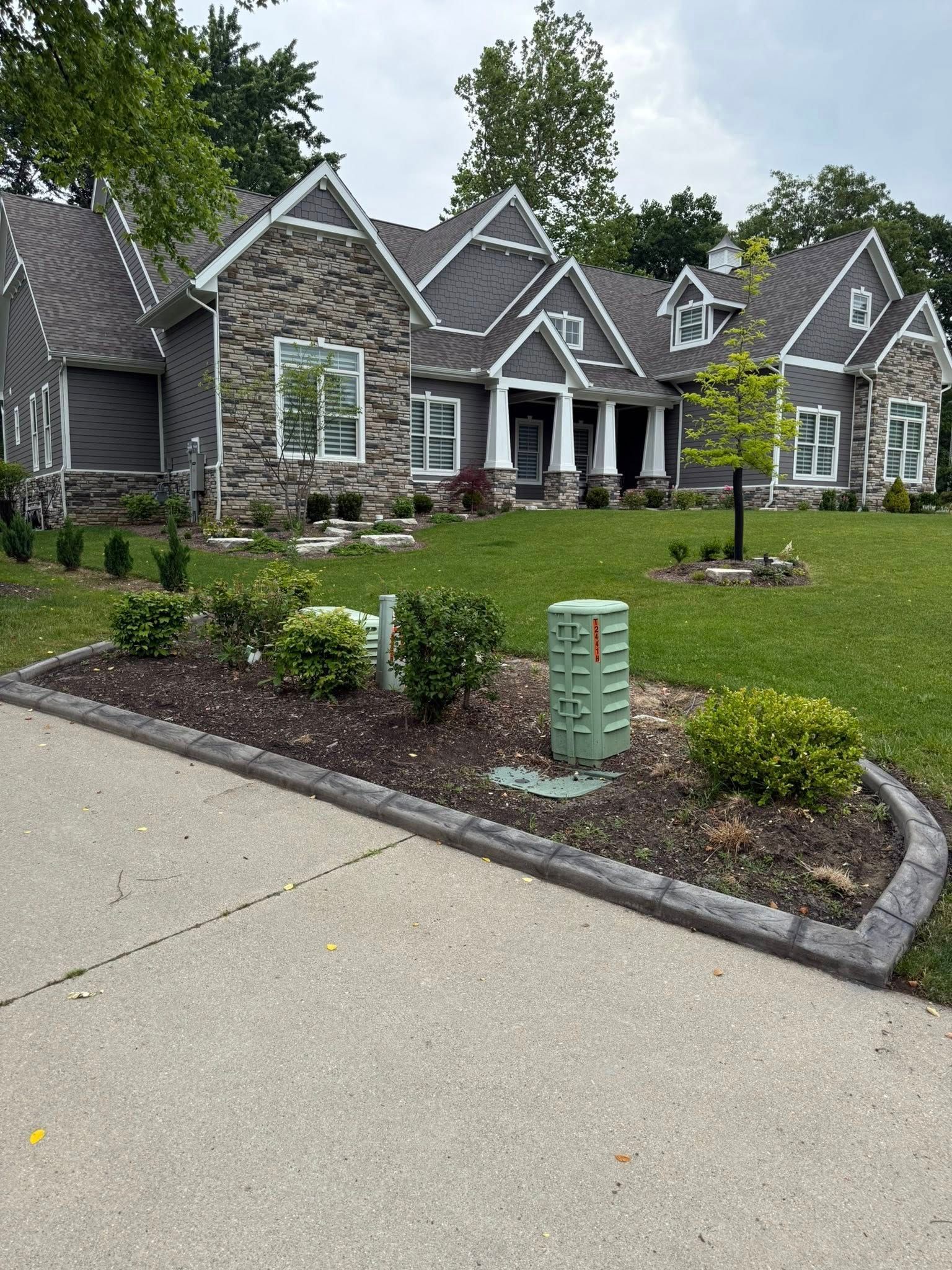 A single-story gray house with stone siding features a landscaped front yard with a green utility box and a concrete drive.