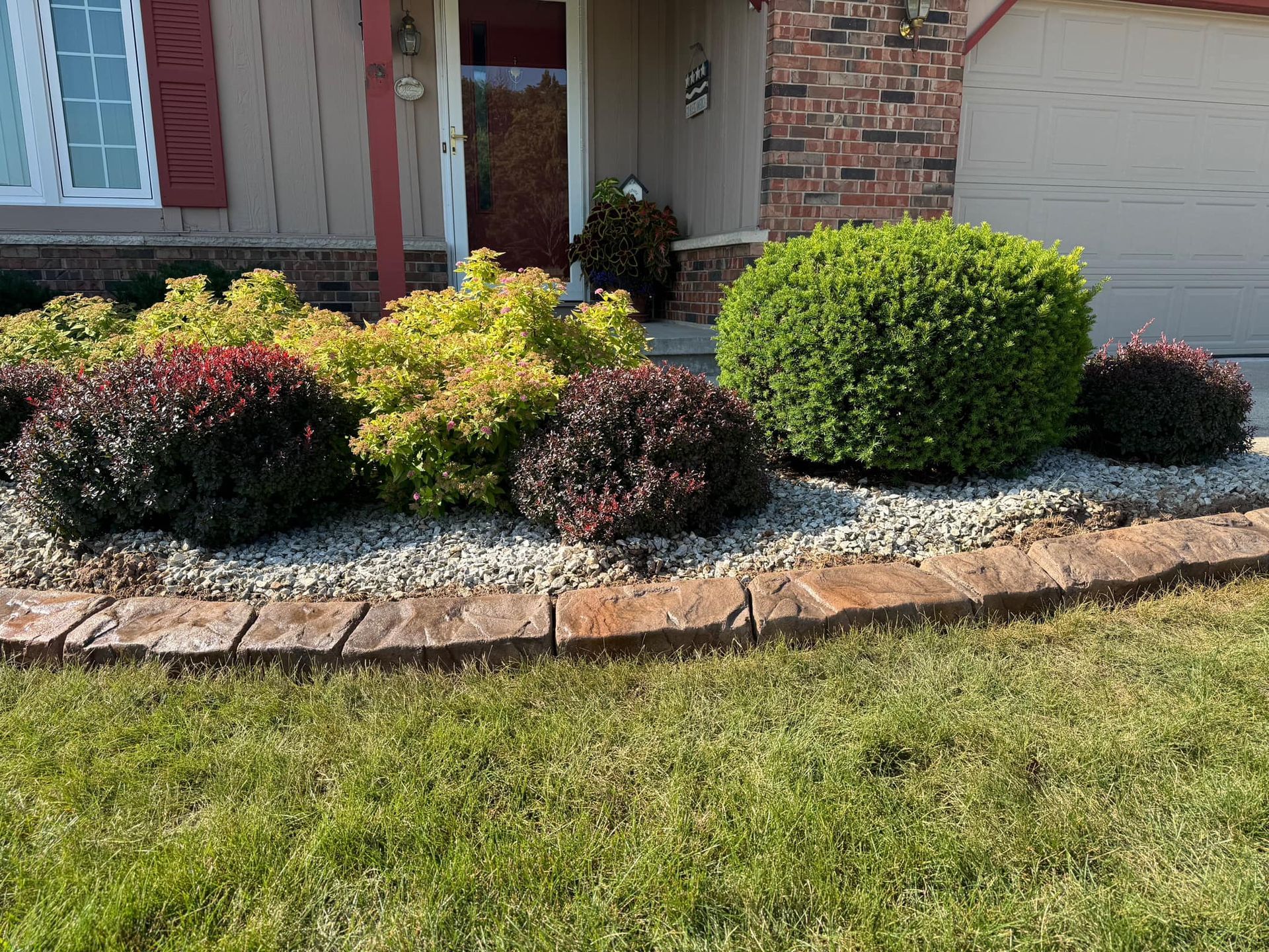 Landscaped garden bed with colorful shrubs and stone edging in front of a house with a brick exterior.