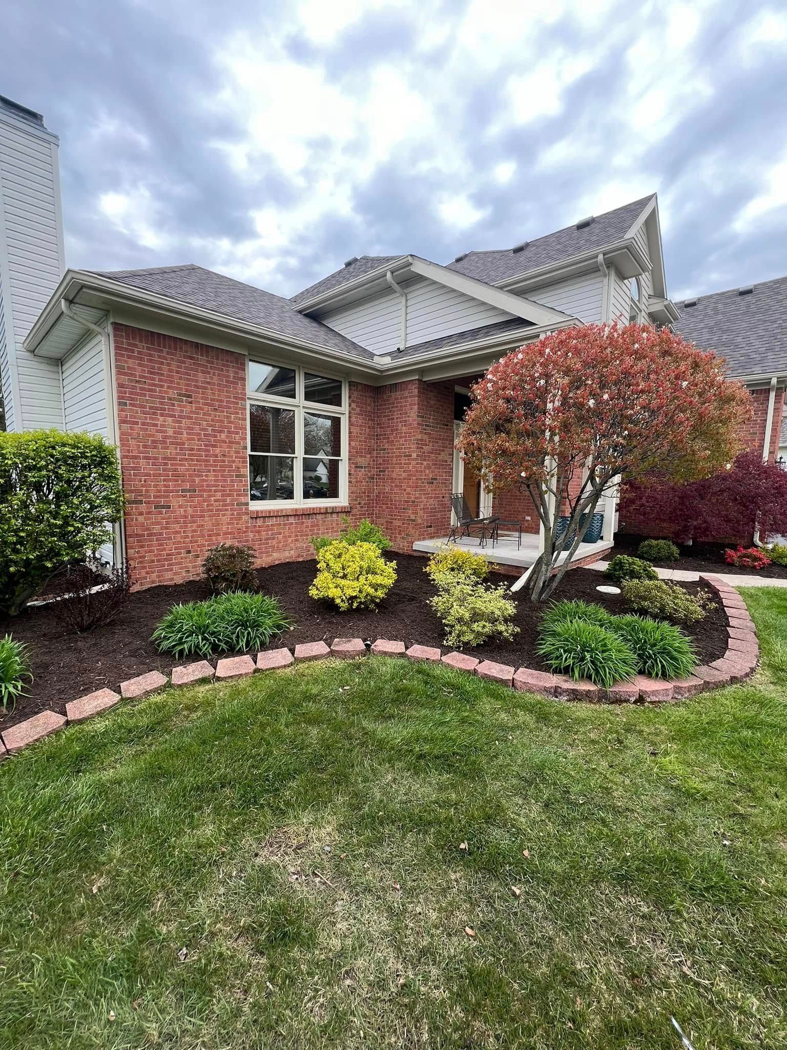 A single-story red brick house with a dark shingled roof, landscaping, and a trimmed round tree under a cloudy sky.