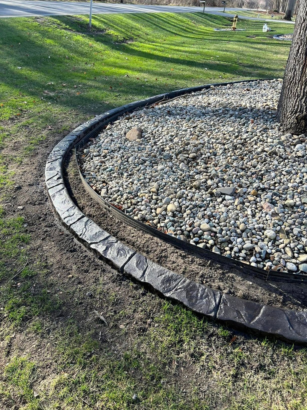 A curved, dark grey, textured stone garden border surrounding a bed of river rocks beneath a tree on a grassy lawn.