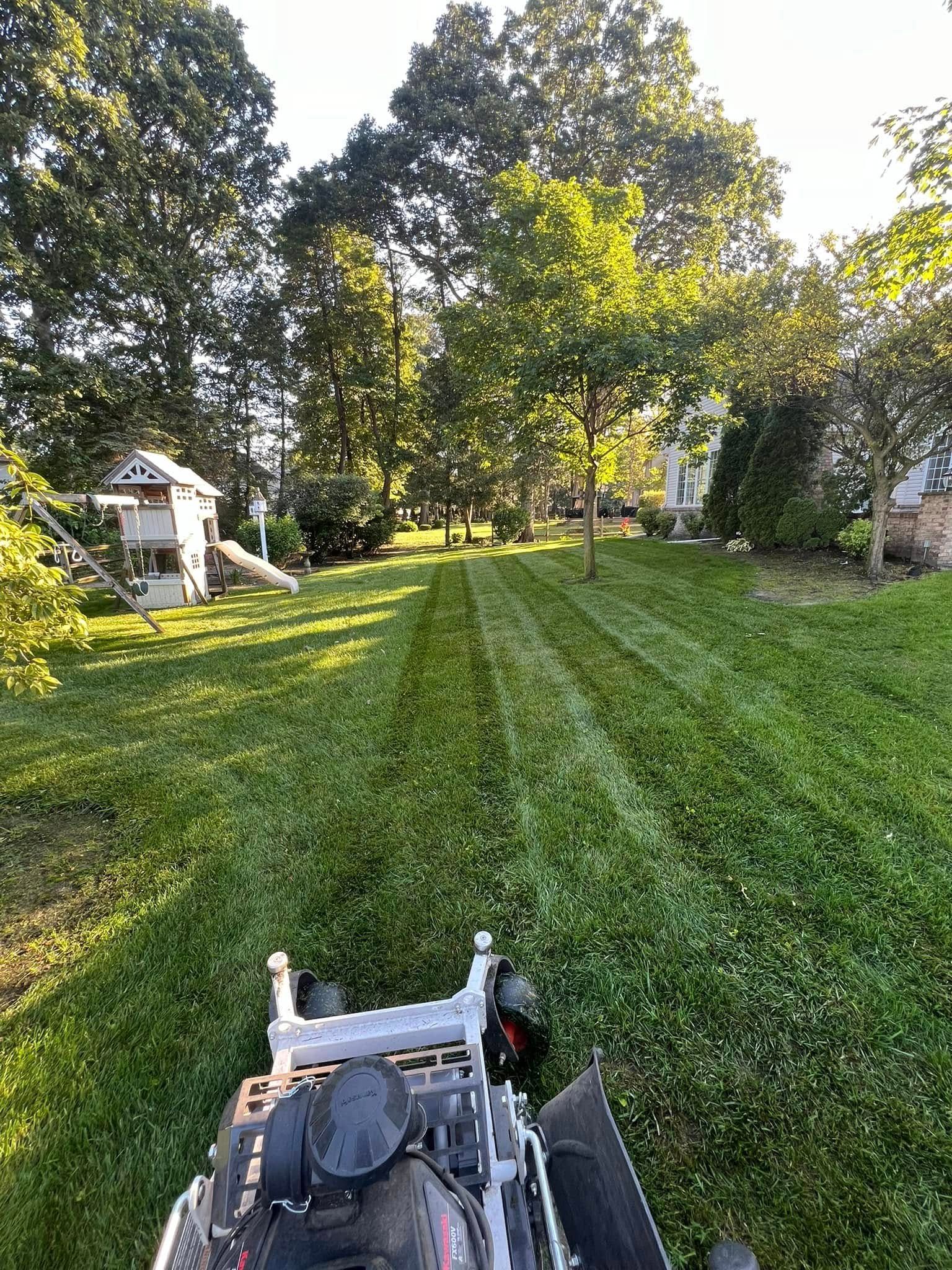 A view from a zero-turn mower cutting grass in a sunny, green backyard with a small playhouse in the distance.