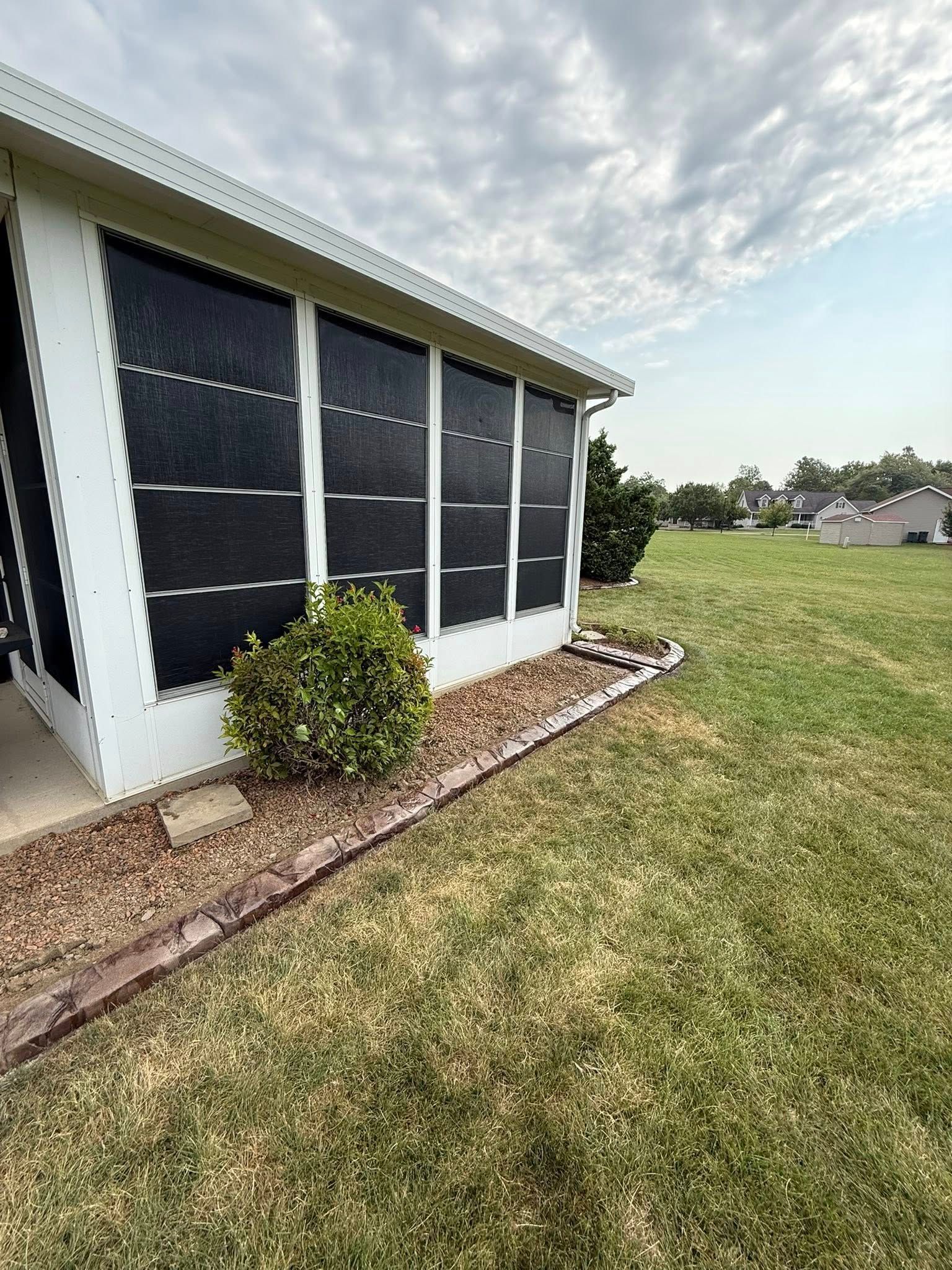 A white sunroom exterior featuring dark solar screens, a small shrub, and a decorative stone border next to a grass lawn.