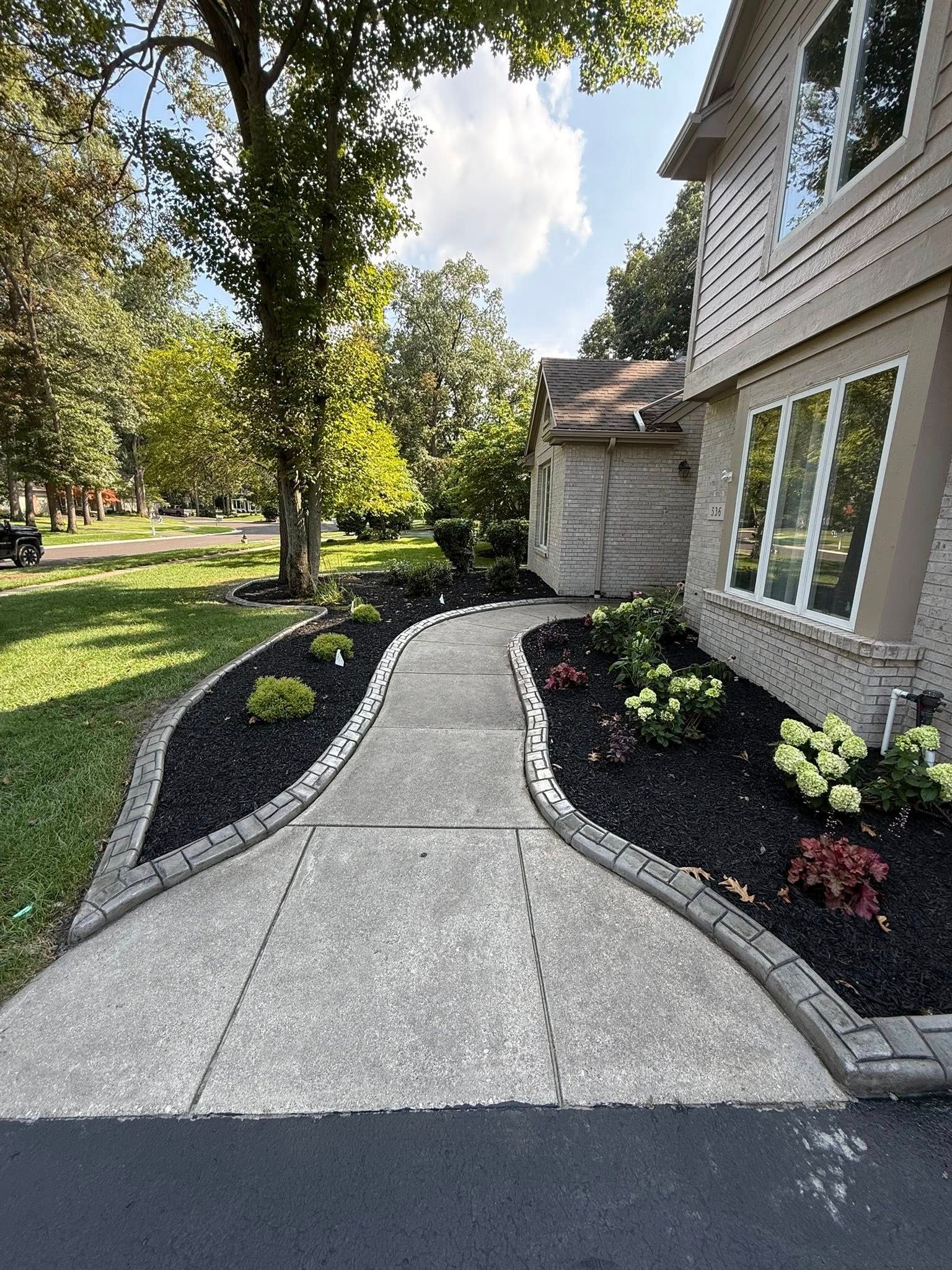 A concrete walkway curves toward a two-story home, bordered by dark mulch and manicured landscaping in a sunny yard.