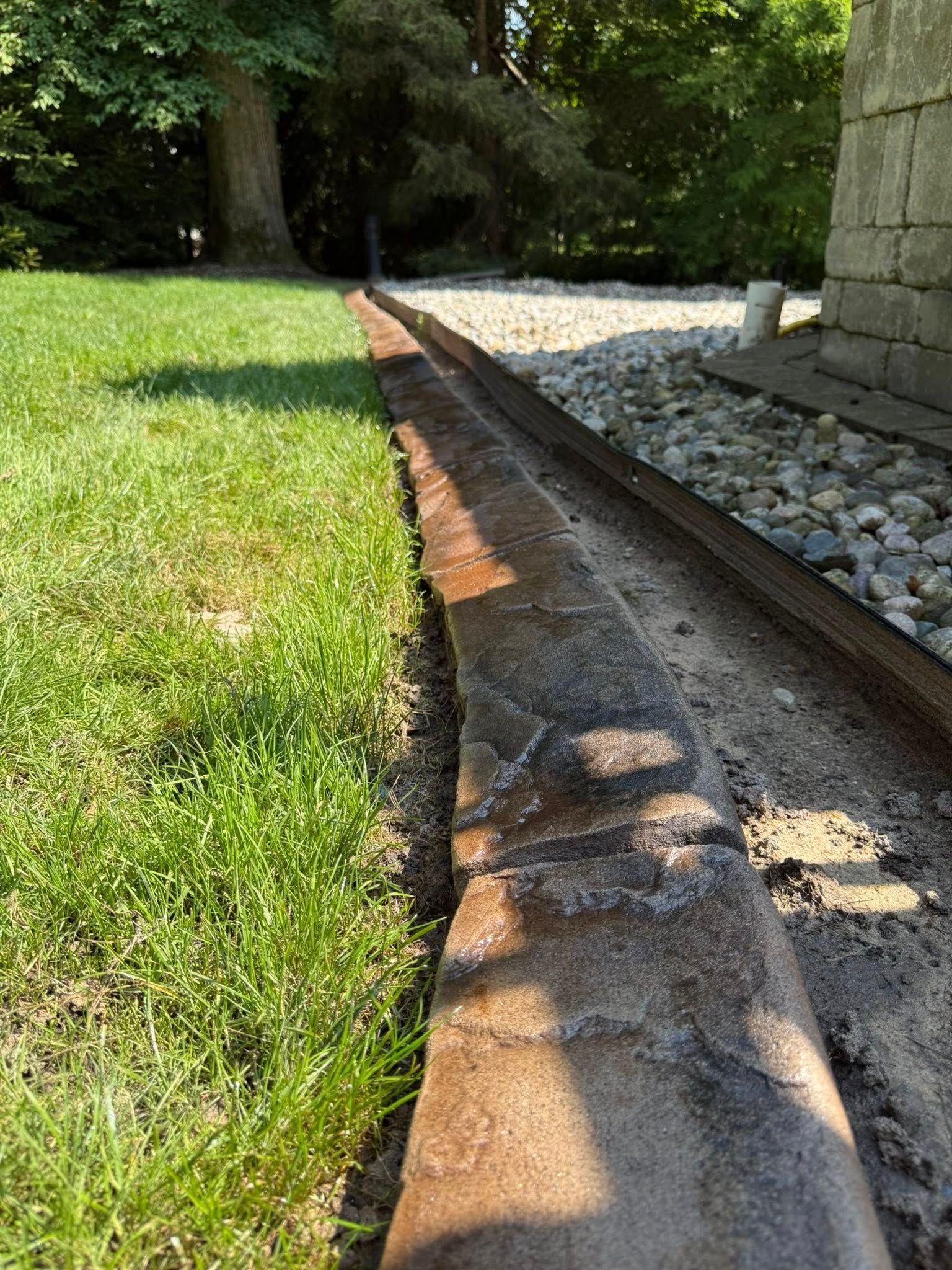 A narrow concrete landscape border separating a green grass lawn from a gravel path next to a stone wall.