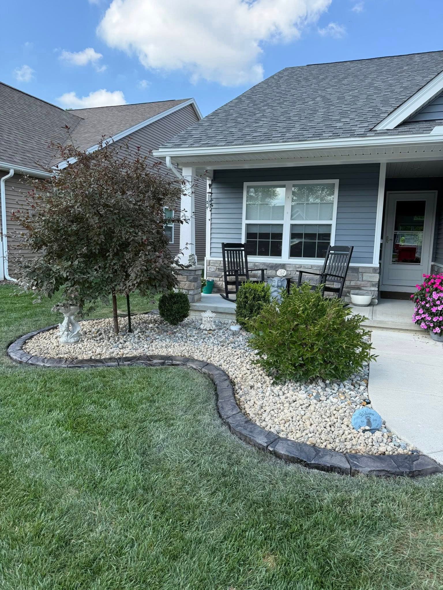 A landscaped garden bed with decorative gray stone curbing, light-colored gravel, a small tree, and green shrubs.