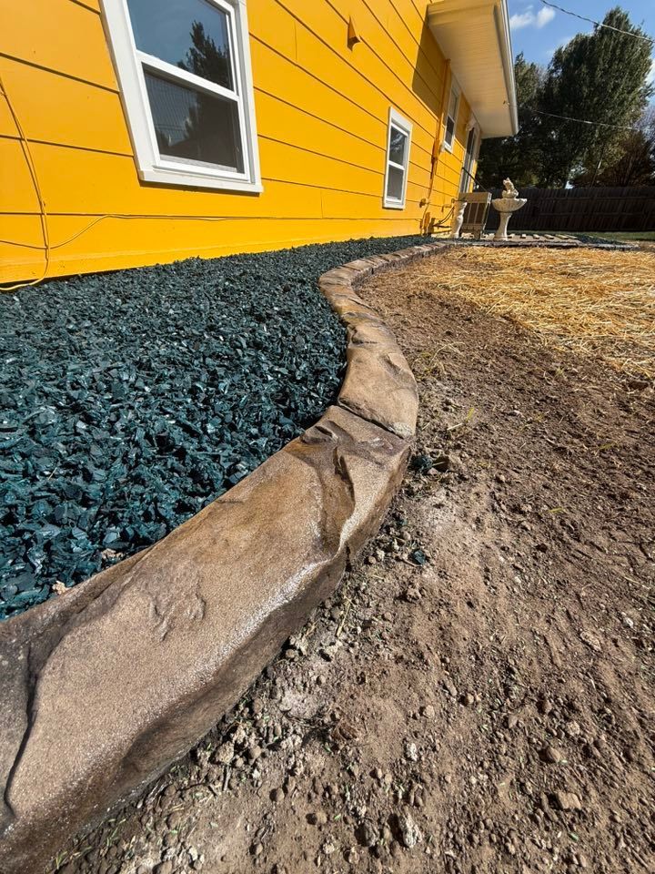 A low, stamped-concrete stone border separates blue gravel landscaping from a dirt yard next to a bright yellow house.