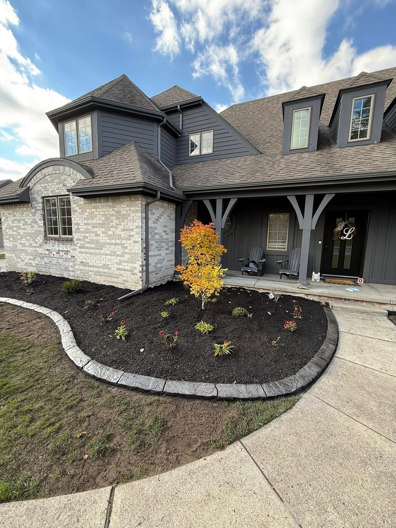 A stone-front house with dark grey roofing and siding, a covered porch, and a landscaped bed with a central autumn tree.