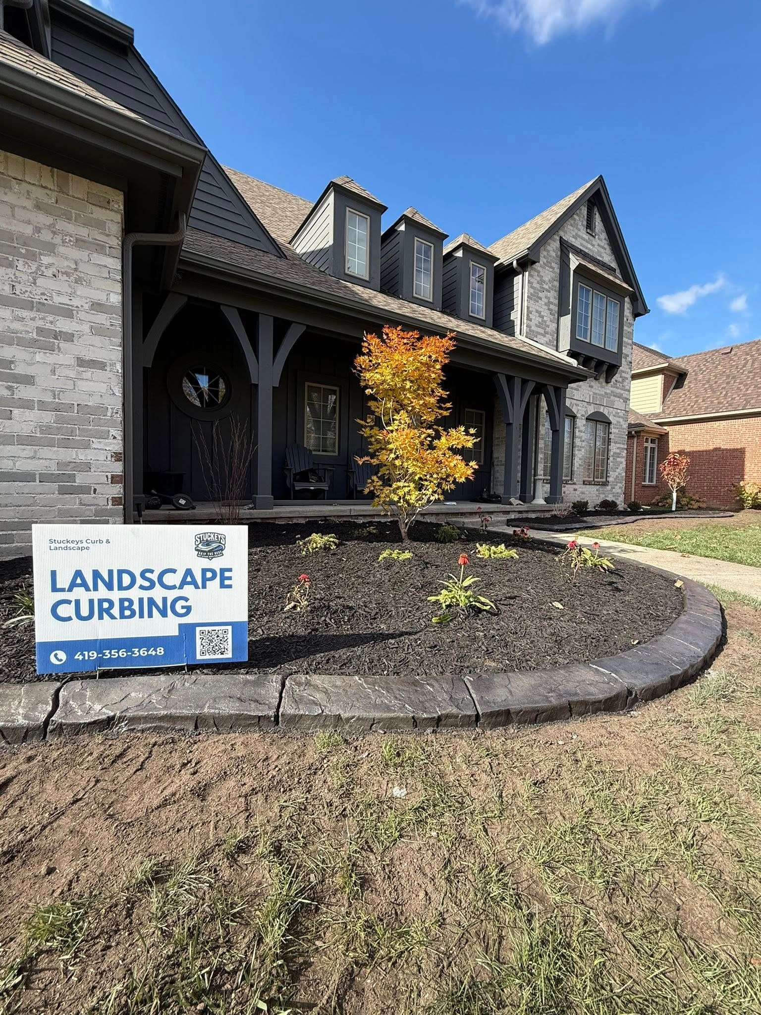 A front yard with new dark gray stone landscape curbing installed around a flower bed in front of a brick house.