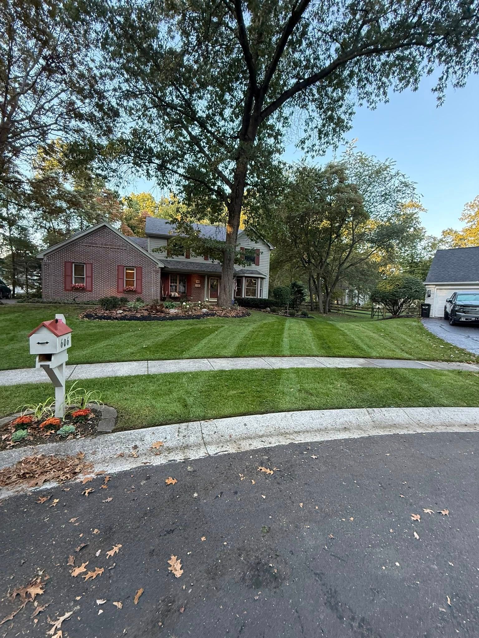 A suburban stone-faced house with a neatly mown lawn, a mailbox in the foreground, and a large tree under a clear blue sky.