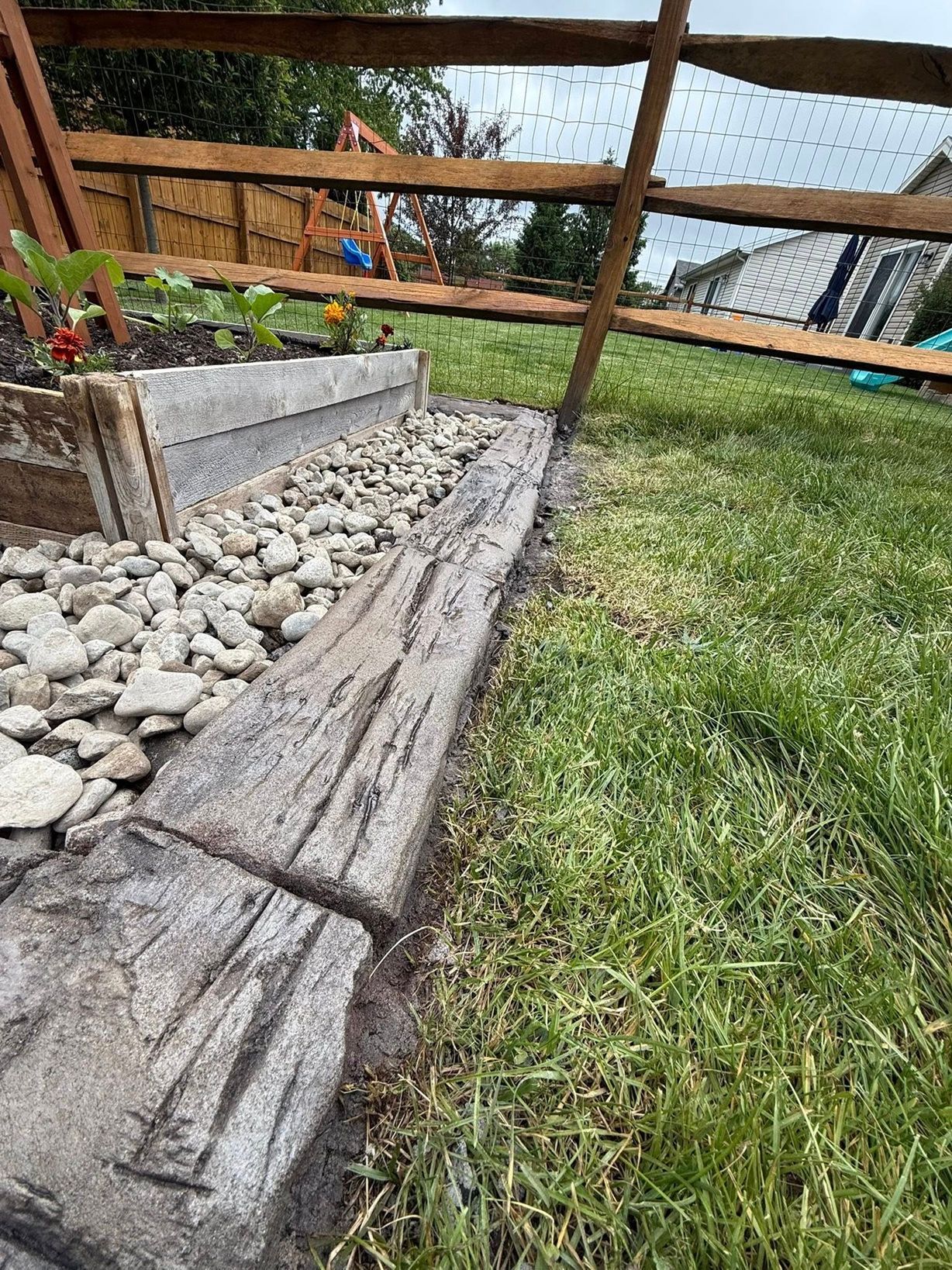 A close-up view of a garden bed bordered by textured concrete timbers and white landscape stones against a grass lawn.