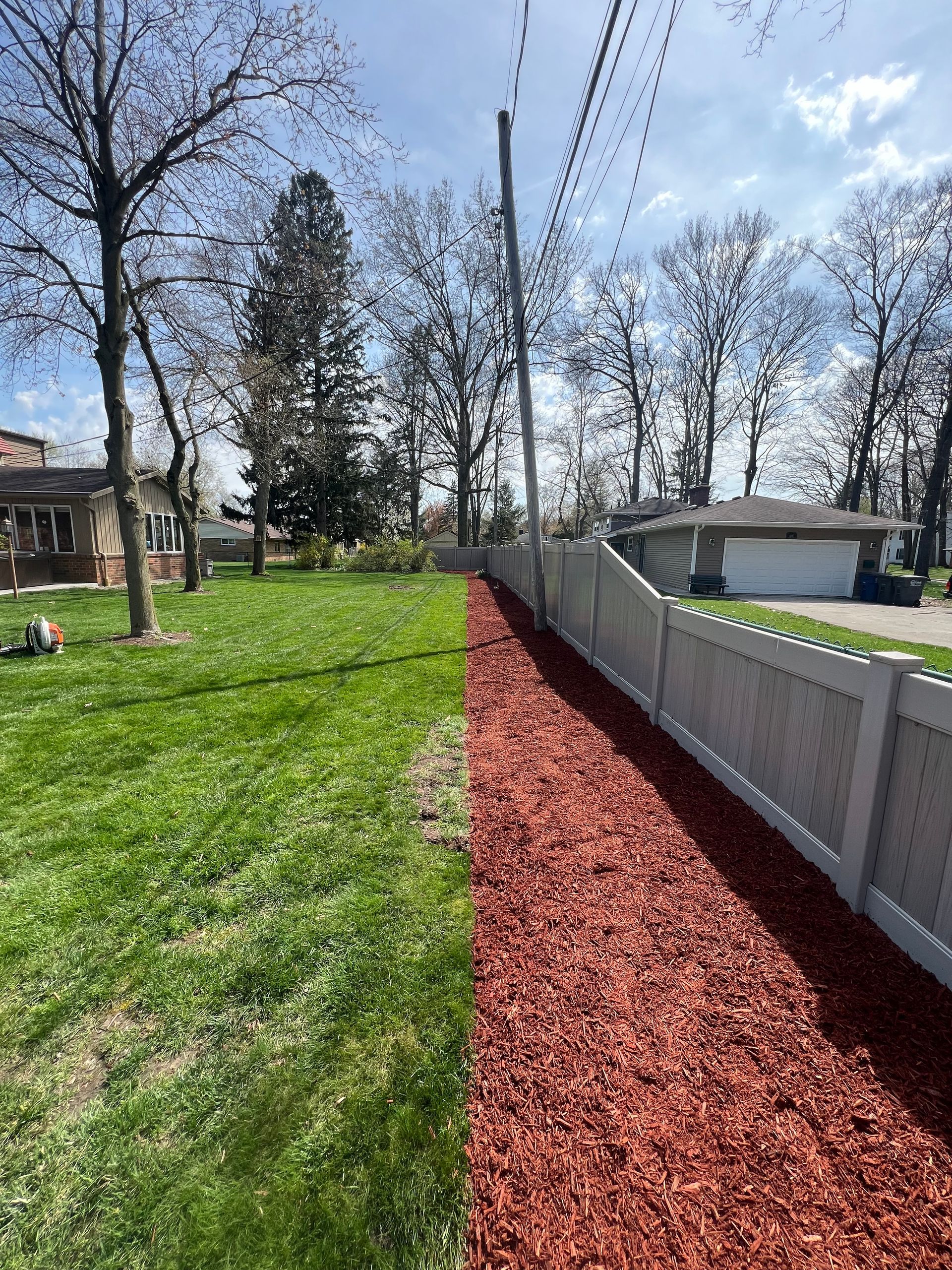 A white vinyl fence runs along a strip of red mulch next to a green grass lawn, with trees and houses in the background.