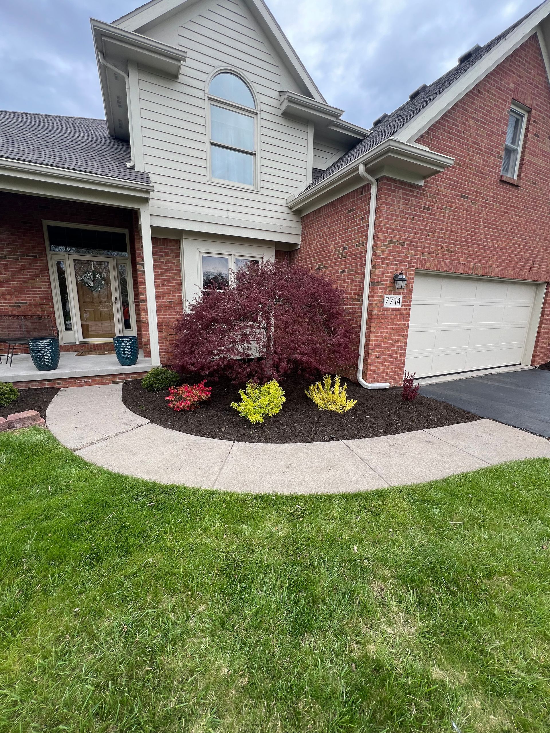 A two-story house with brick and siding, a curved walkway, a large red bush, and two smaller yellow shrubs in mulch.