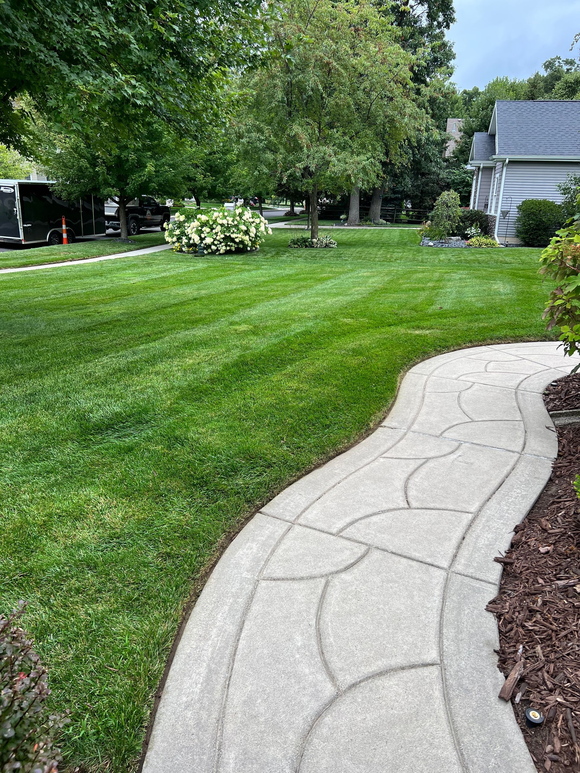 A curved concrete path with a decorative arched pattern winds through a lush green residential lawn toward a house.
