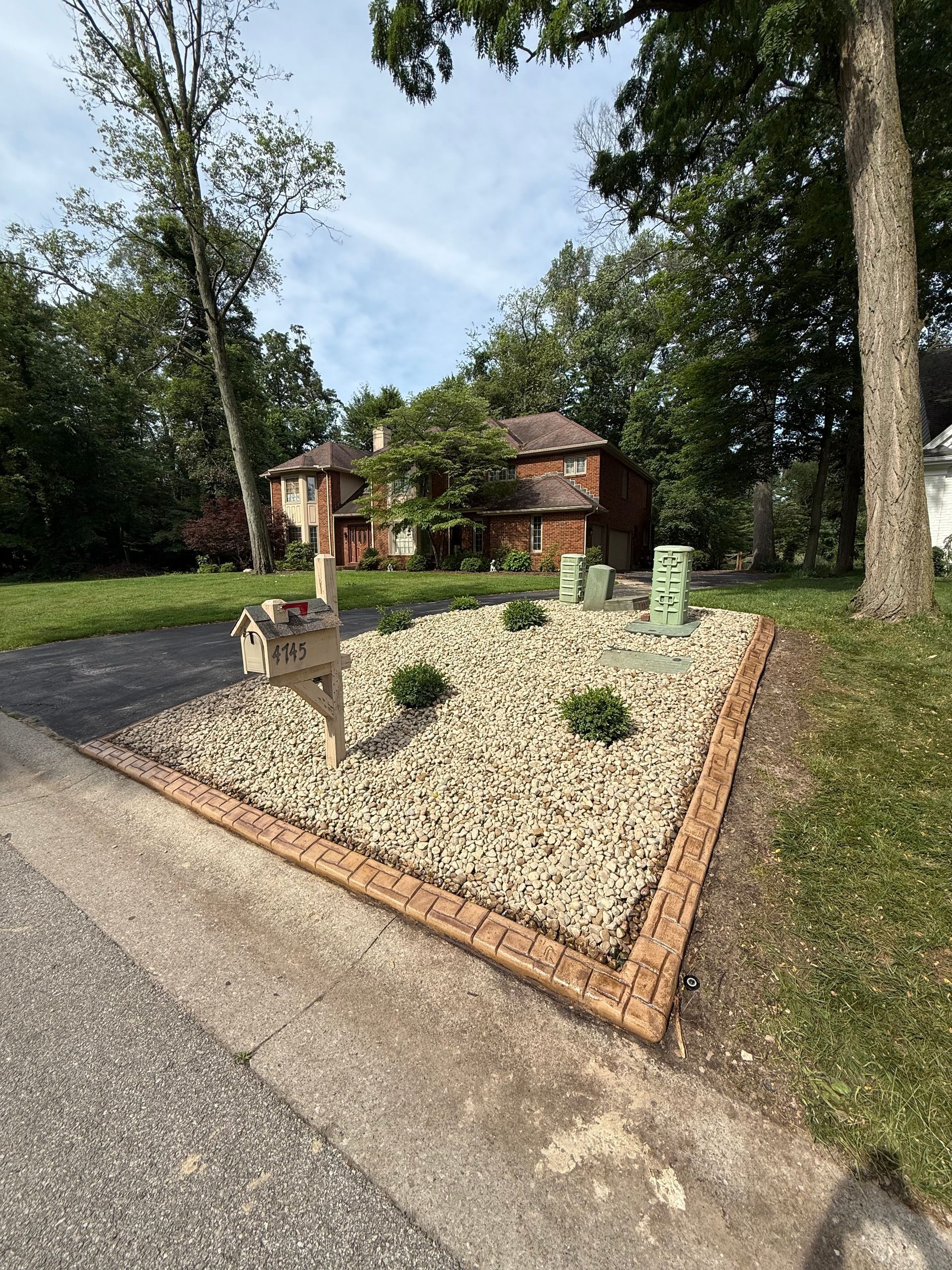 A gravel garden bed near a driveway with a wooden mailbox, decorative stones, small shrubs, and two small grave markers.