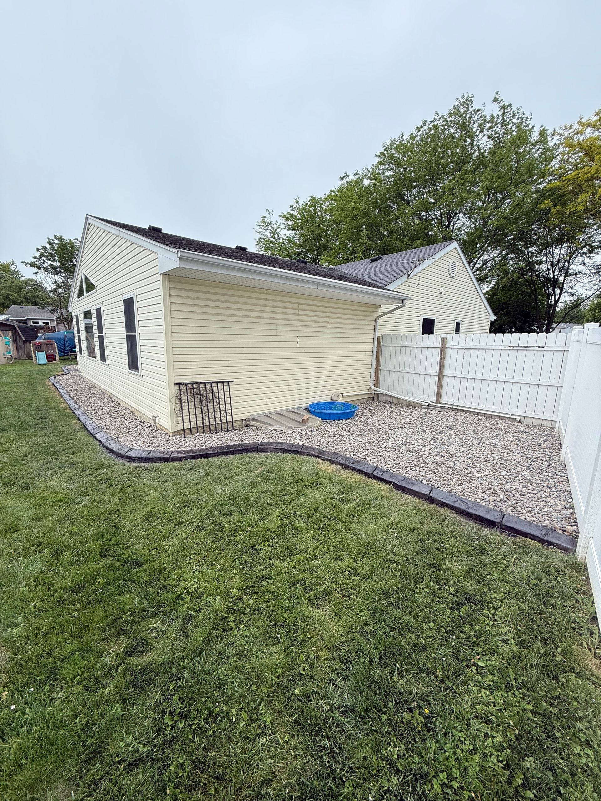 A side view of a light yellow house with stone landscaping, a blue pool toy, and a white privacy fence against a lawn.