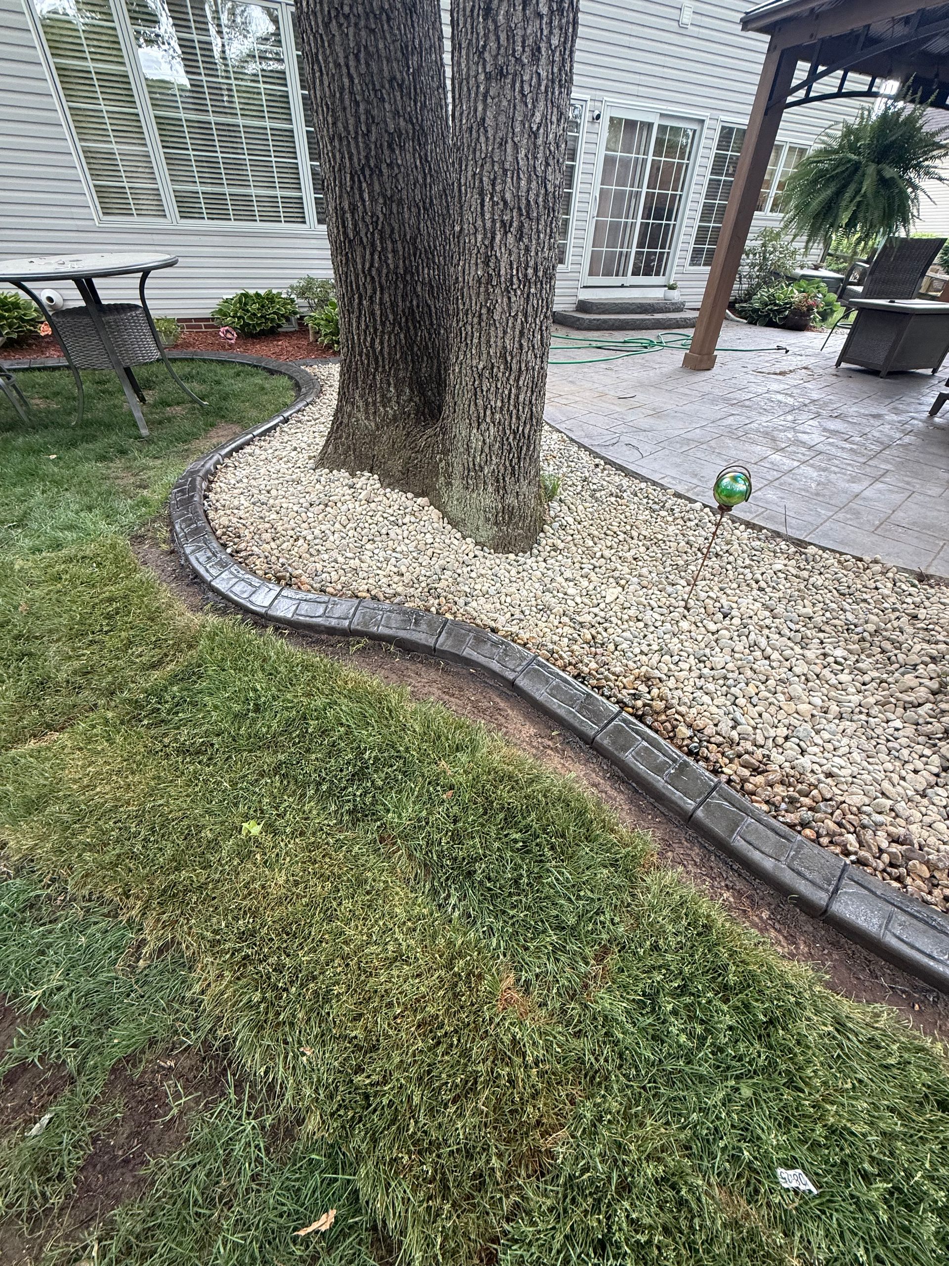 A curved concrete border separates a green lawn from a gravel landscaping bed surrounding a tree near a patio.