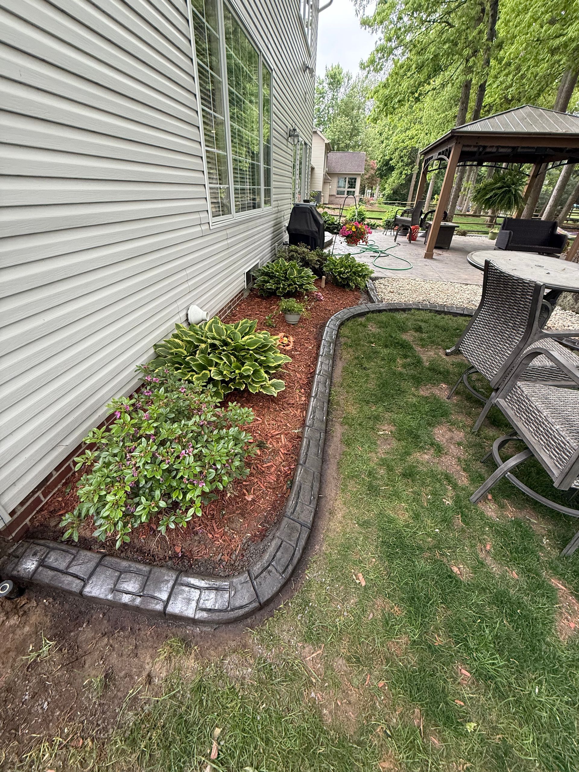 A curved, stone-look garden border separates a mulch-filled flower bed from a grassy lawn next to a vinyl-sided house.