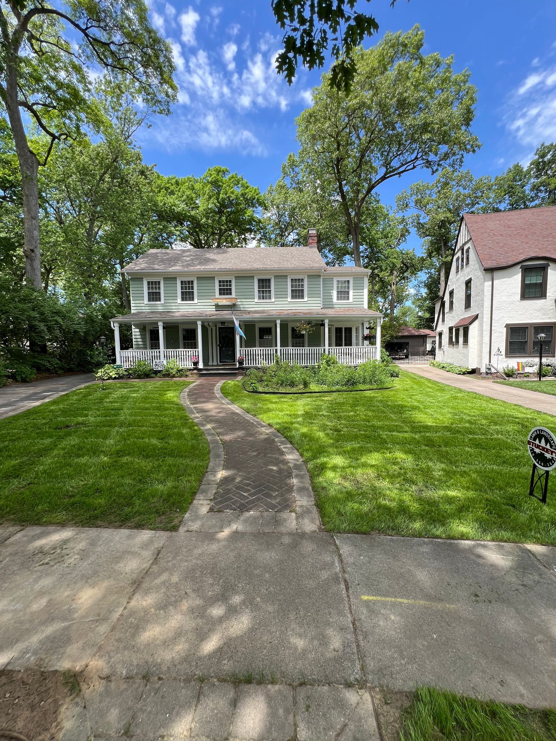 A light-green two-story house with a white front porch, surrounded by lush green lawns and trees under a blue sky.