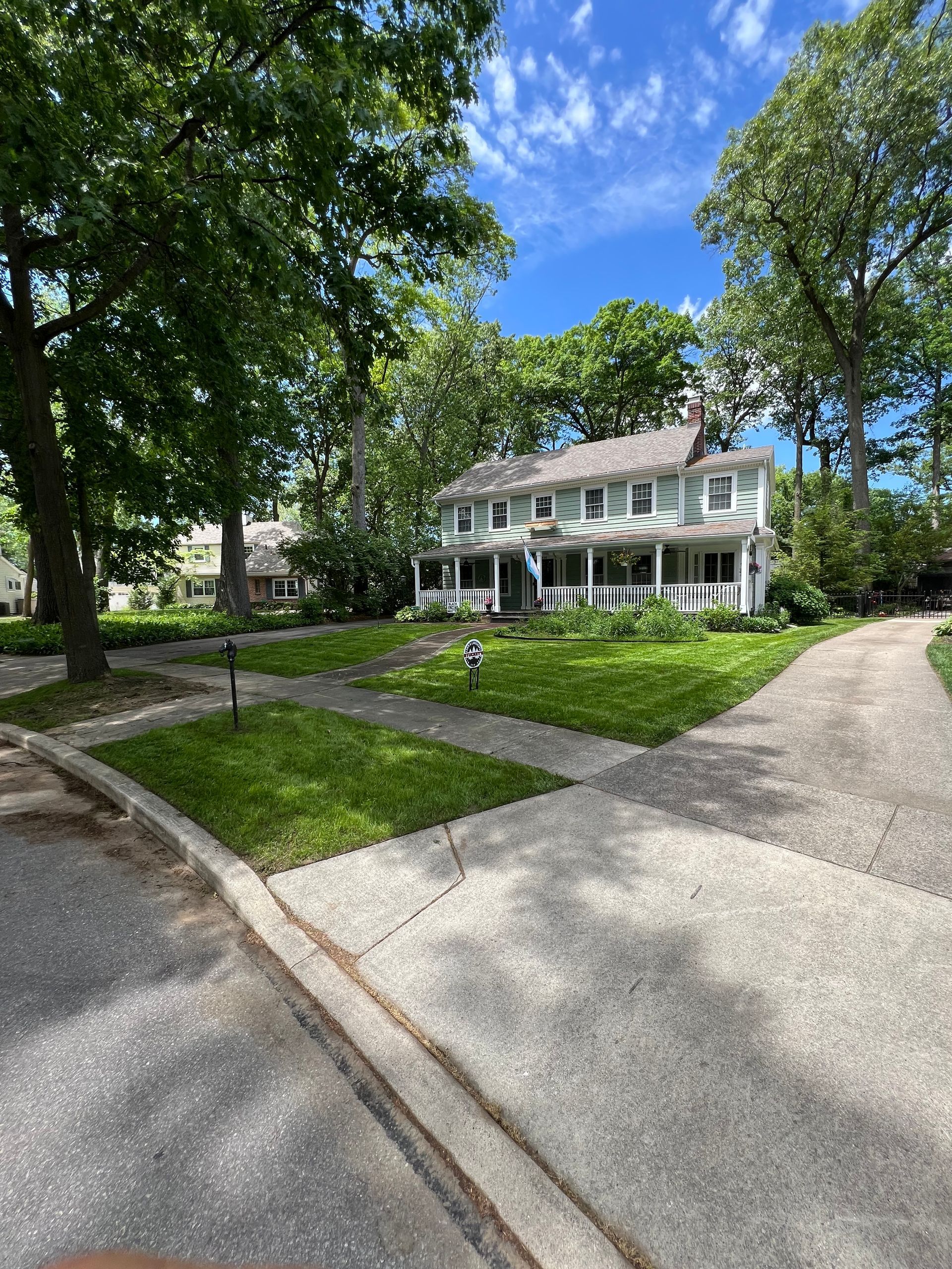 A two-story white house with a porch and green shutters sits behind a sunny front lawn surrounded by large trees.