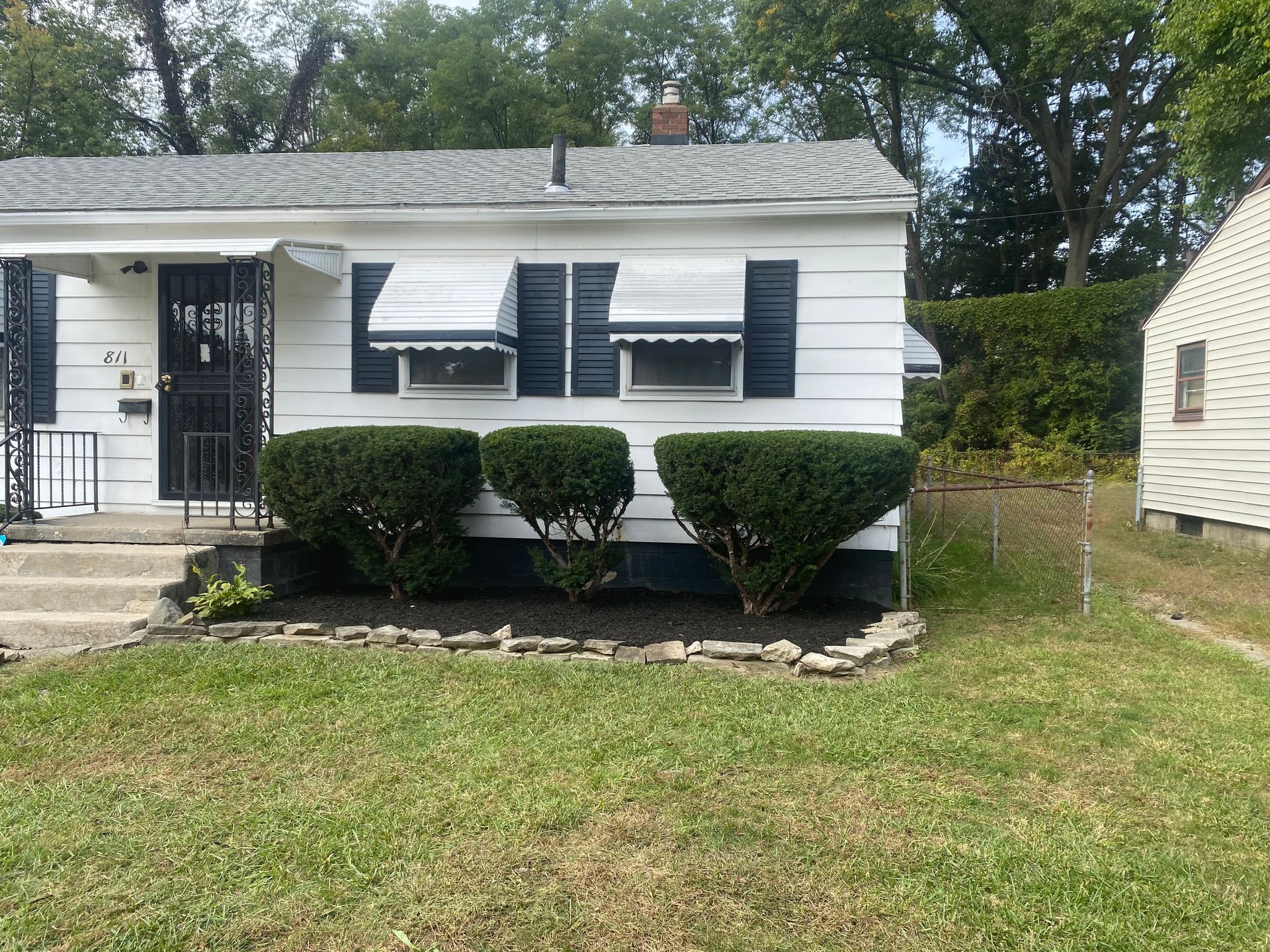 A white one-story house with dark shutters, striped awnings, and three rounded shrubs in a mulched bed.